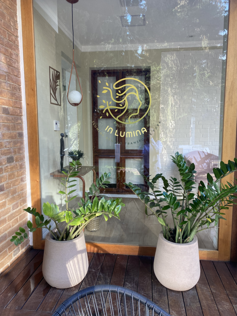 Entrada do espaço presencial do In Lumina, com o logotipo da marca na porta de vidro e plantas naturais, em São José dos Campos